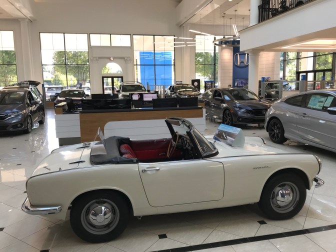 White Honda S800 convertible on showroom floor with newer Hondas in background.