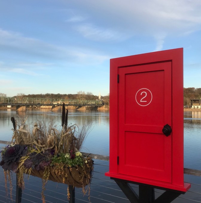 Small red door, with a number 2 on it, beside river. A pot of plants sits beside it.