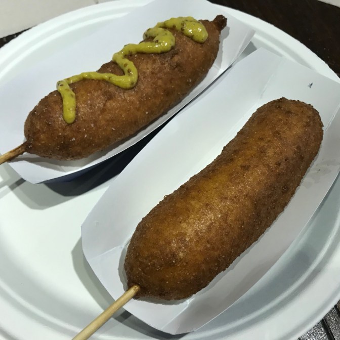 Two corn dogs, one with mustard, on a paper plate.