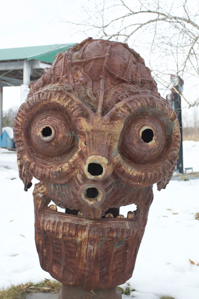 Metal sculpture of welder’s helmet on head. Large goggles dominate the sculpture.