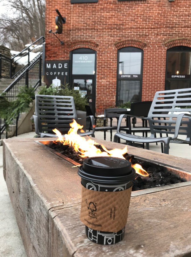 Cup of coffee on side of outdoor fire pit, with brick coffee shop in background.