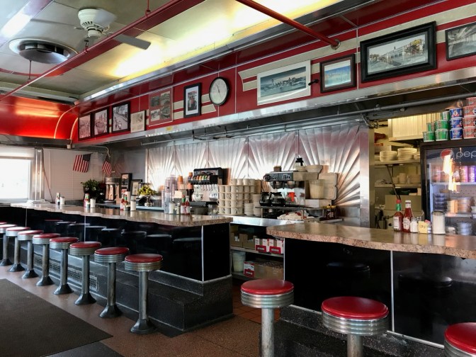 Interior of diner, including two counters with seating for twelve, and an open doorway looking into the kitchen.