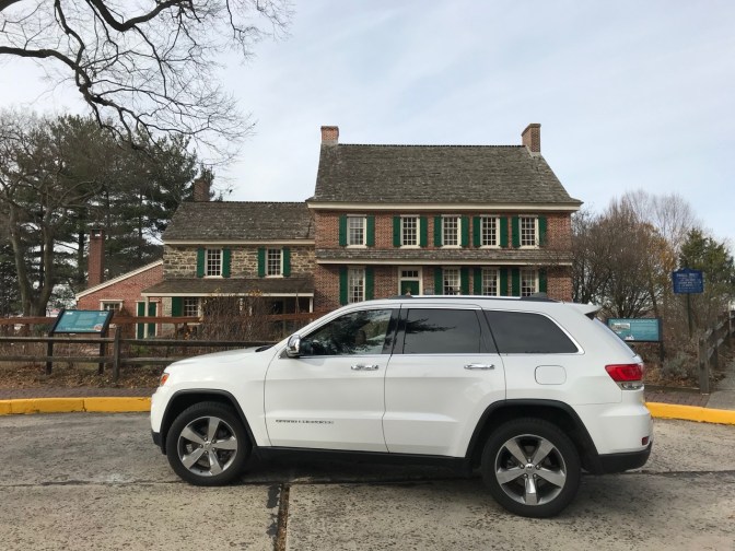 White Jeep Grand Cherokee parked in front of two-story brick colonial-era house.