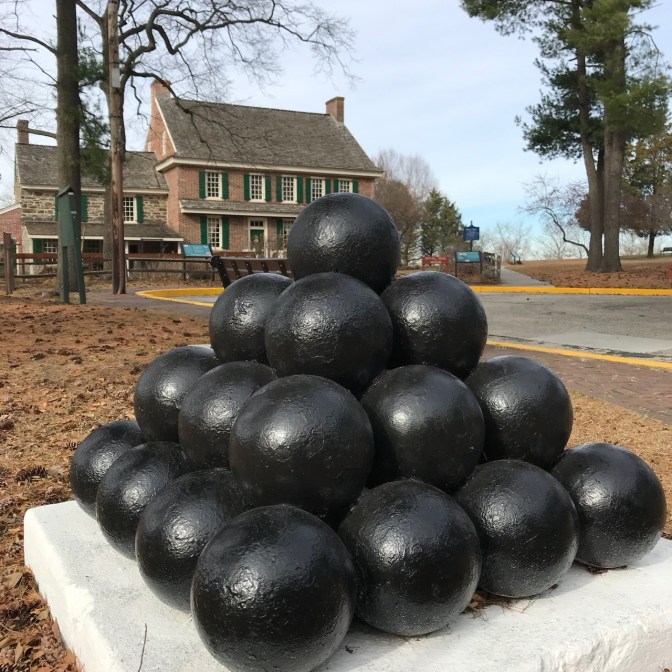 Pyramid of cannon balls, with red brick two-story house in background.
