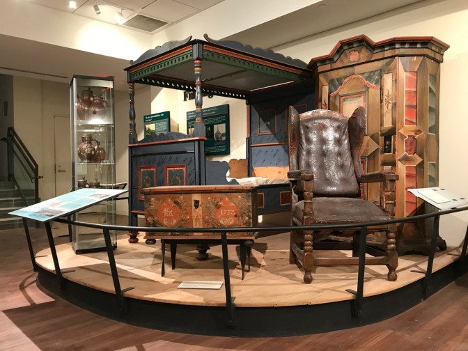 Museum display of bedroom furniture including a four poster-bed, a chest, a chair, and an armoir. 