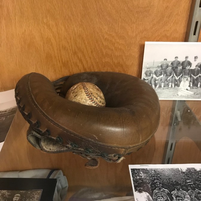 Catcher’s mitt on glass shelf, with photos of old baseball teams surrounding it.