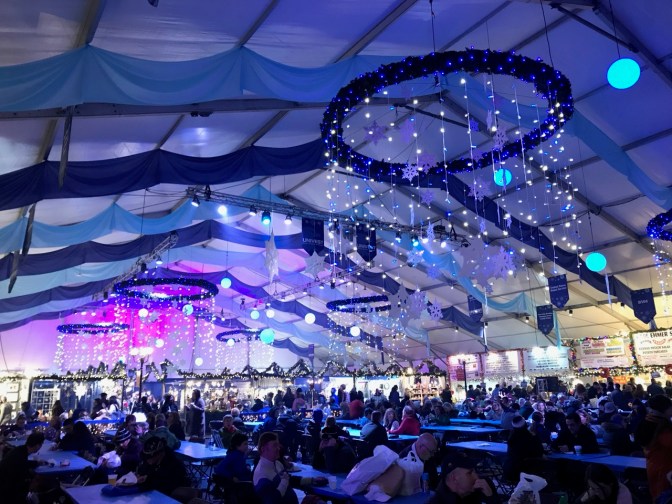 Interior of one of the main tents of the Christmas Market, with people seated at tables in foreground, shops in the background, and snowflake chandeliers hanging from ceiling.