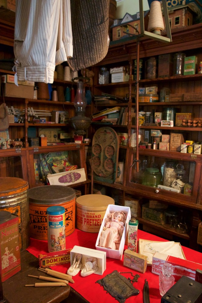 Shelves and counter of general store, with items such as cans, jars, dolls, and toys.