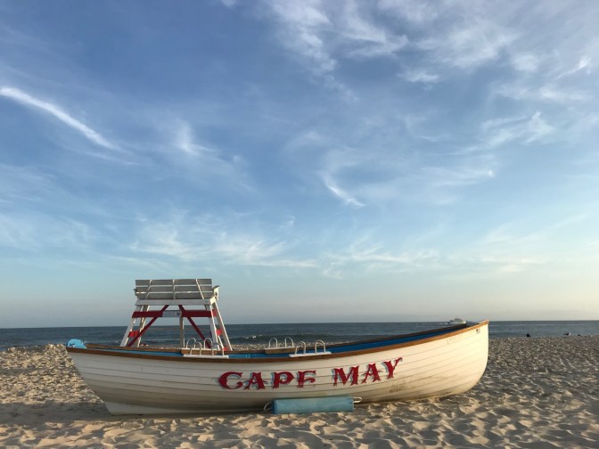 Row boat that says CAPE MAY on beach, in front of life guard stand.