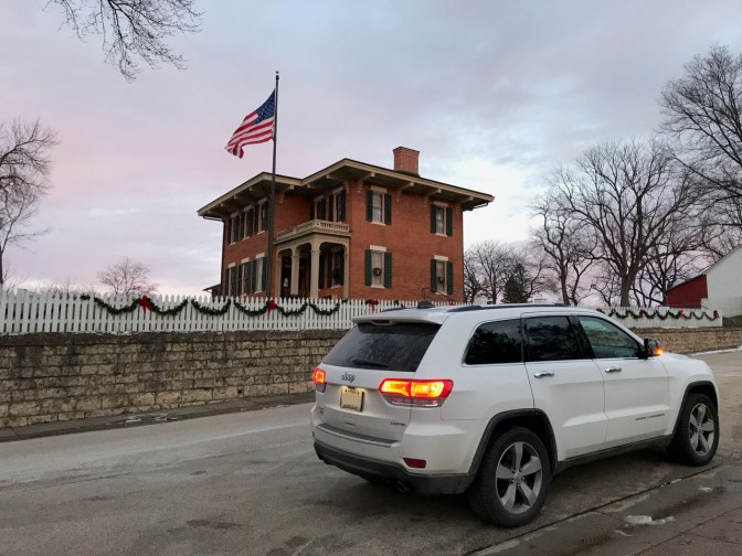 White Jeep Grand Cherokee parked in front of brick house, with a large American flag flying in the distance.