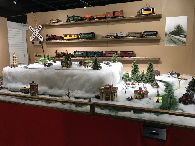 Room filled with a table with a model train set, and model trains on the walls.