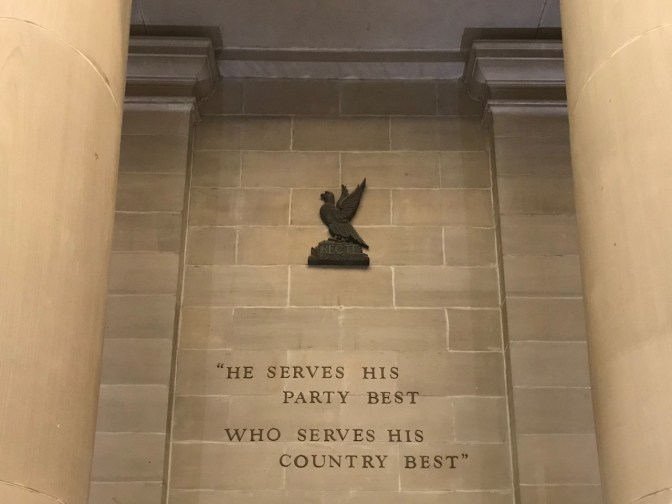 Text on wall of atrium that says HE SERVES HI PARTY BEST WHO SERVES HIS COUNTRY BEST.