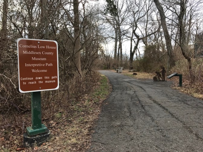 Path in woods. A sign beside it says CORNELIUS LOW HOUSE - MIDDLESEX COUNTY MUSEUM INTERPRETIVE PATH WELCOME CONTINUE DOWN THIS PATH TO REACH THE MUSEUM