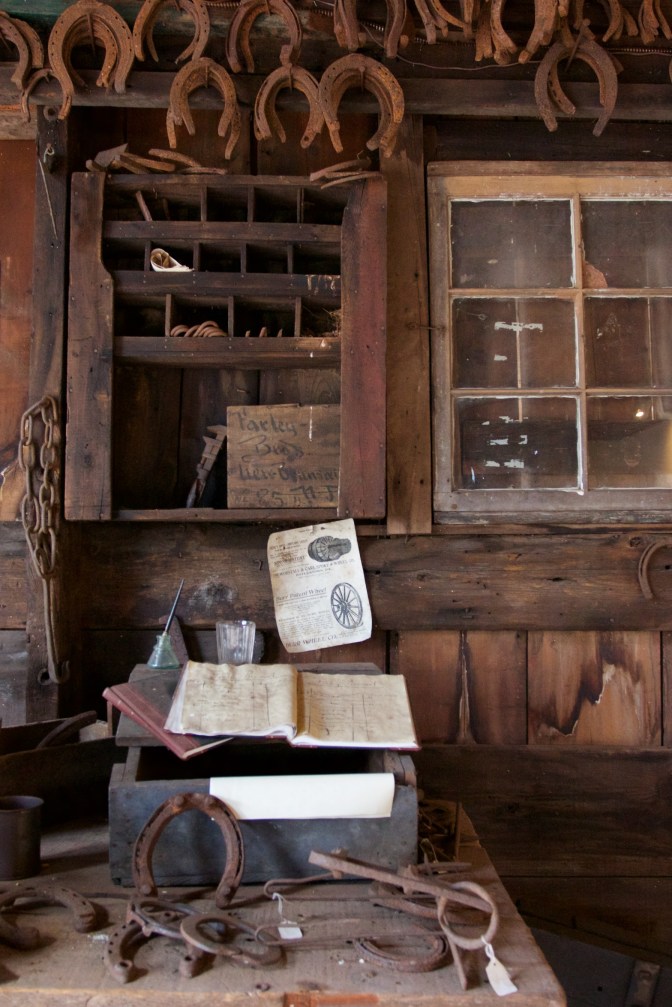 Interior of blacksmith shop. A table on foreground has ledgers and horseshoes. A cabinet with equipment is on the walls, and horseshoes hang from the ceiling.