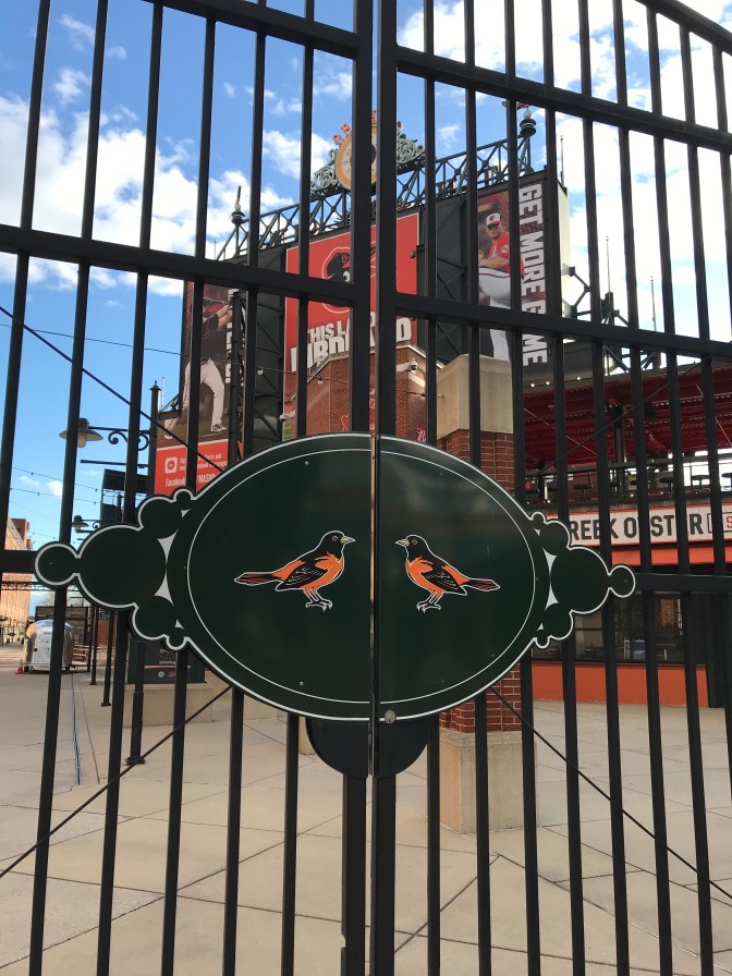 Gate with Orioles on latch of gate, with Camden Yards stadium in background.