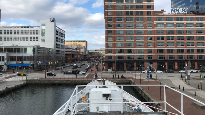 View of forecastle of ship and main gun, with Baltimore skyline behind it.