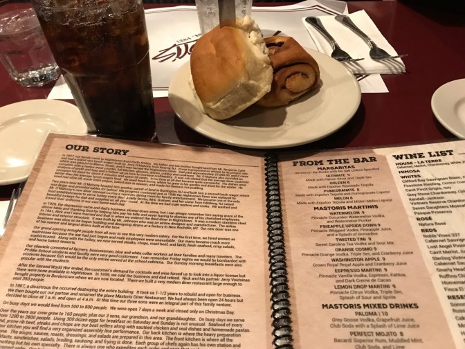 Menu from Mastoris, open to "Our Story." Two rolls are on the table, along with beverages.