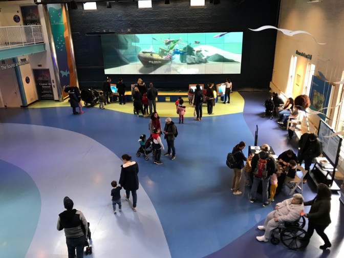 View of lobby of Aquarium, with multiple families walking through, some watching a large video screen on far wall.