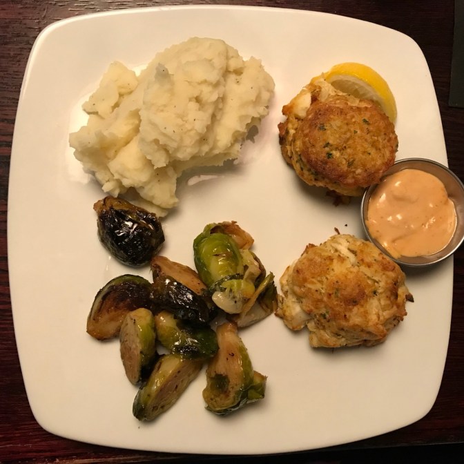White plate with crab cakes, Brussels sprouts, and mashed potatoes.