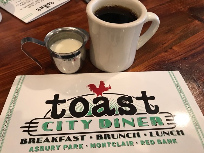 Coffee in white mug and cream in silver pitcher, in front of menu that says TOAST CITY DINER BREAKFAST BRUNCH LUNCH