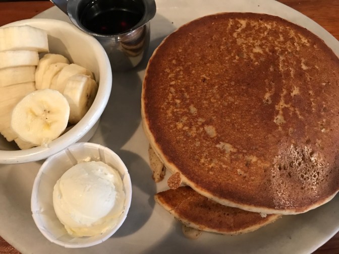 Pancakes, small container of butter, small pitcher of maple syrup, and bananas on a white plate.