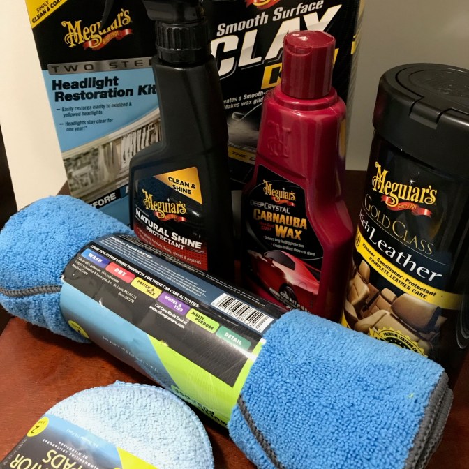 Table covered with car care products including waxing pads, microfiber cloths, Meguiar's Leather Care, Meguiar's Carnuba Wax, Meguiar's Natural Shine protectant, Meguiar's Smooth Surface Clay Bar, and Meguiar's Two-Step Headlight Restoration.