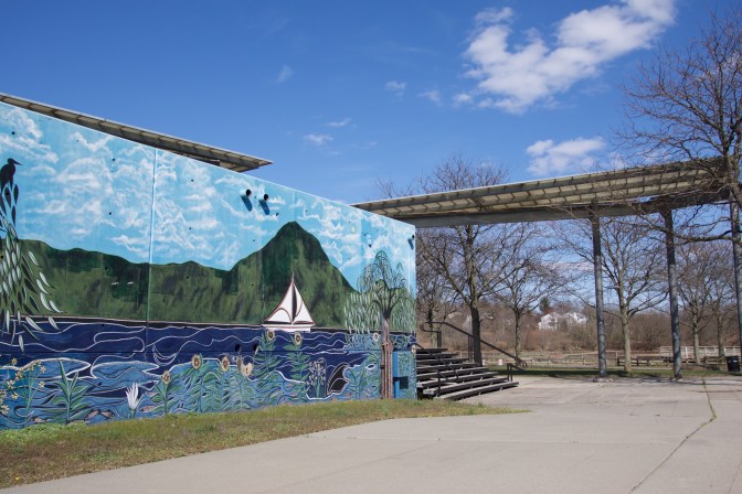 Mural in Body Park of Raritan River, with a boat on the river and hills in the distance. The mural sits on a wall with a pavilion in the background.
