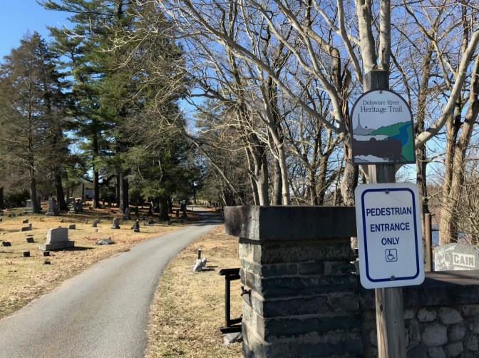 Entrance to cemetery, with sign on right saying DELAWARE RIVER HERITAGE TRAIL and another saying PEDESTRIAN ENTRANCE ONLY.
