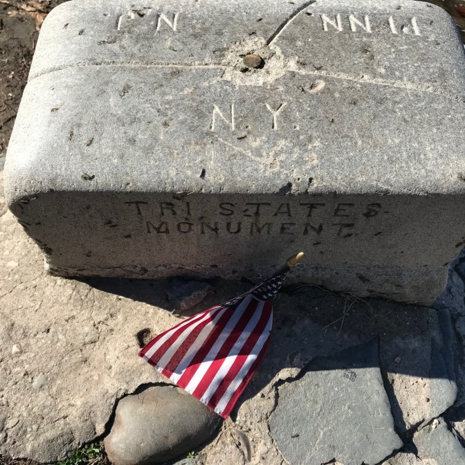 Stone marker with copper nail in middle. Side of marker says TRI STATES MONUMENT. Top of Marker is divided into three sections: NJ, PENN, and NY.