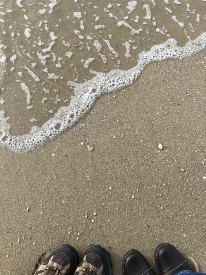 Beach and ocean water, with a man's pair of hiking shoes and a woman's pair of boots in bottom of frame.