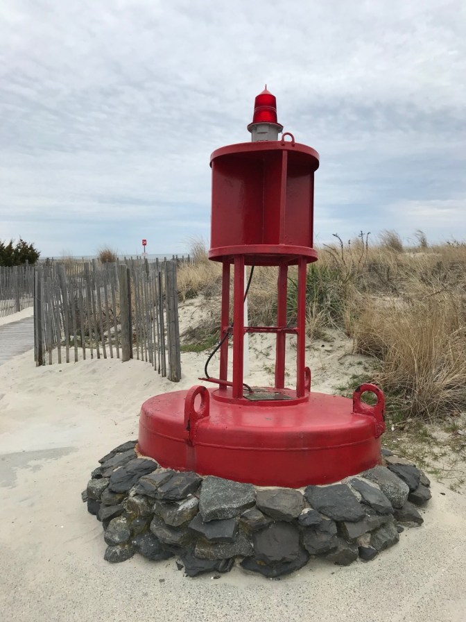 Red Coast Guard Buoy on rocks beside sand dune.