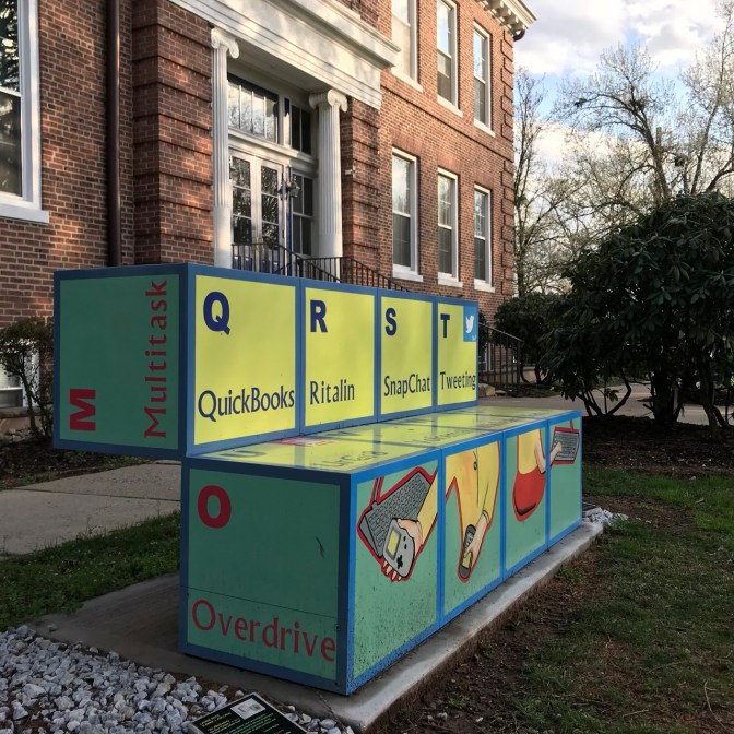 Bench made of large building blocks, with images of technology, and words like Multitask Overdrive Quickbooks Ritalin, SnapChat, Tweeting.
