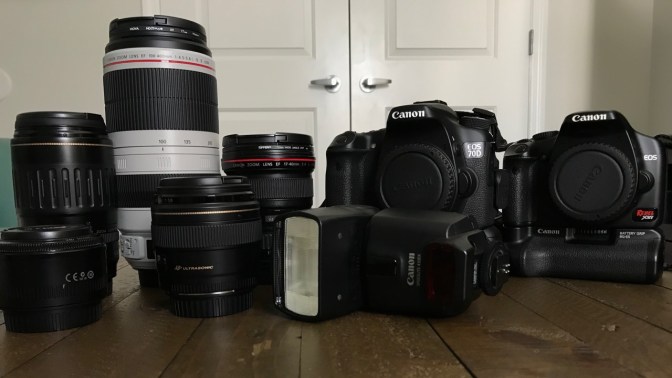 Five camera lenses, one flash, and two camera bodies, on wooden table.