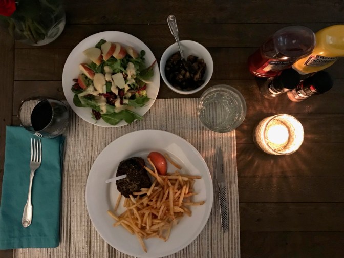 Table with candle, plate with salad, plate with steak and fries, bowl with mushroom, glass of wine, and glass of water.