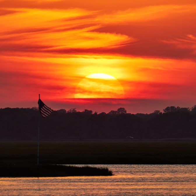 Setting sun over shore, with American flag on pole on beach in foreground.