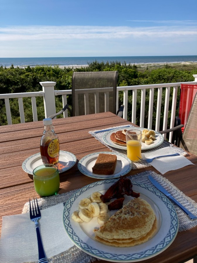 Porch table overlooking ocean. On plates on table are pancakes, bacon, bananas, and scrapple. Two glasses of orange juice are on table as well.