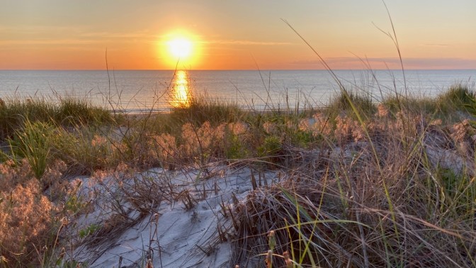 Setting sun over dune in front of ocean.