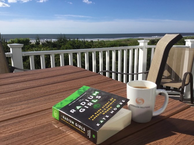 View of ocean from balcony overlooking beach. A cup of coffee and a copy of The Radium Girls by Kate Moore are on a picnic table on deck.