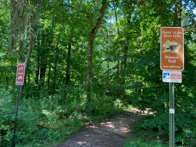 Entrance to hiking trail with sign by right side that says BATTLE OF THE SHORT HILLS HISTORIC TRAIL.