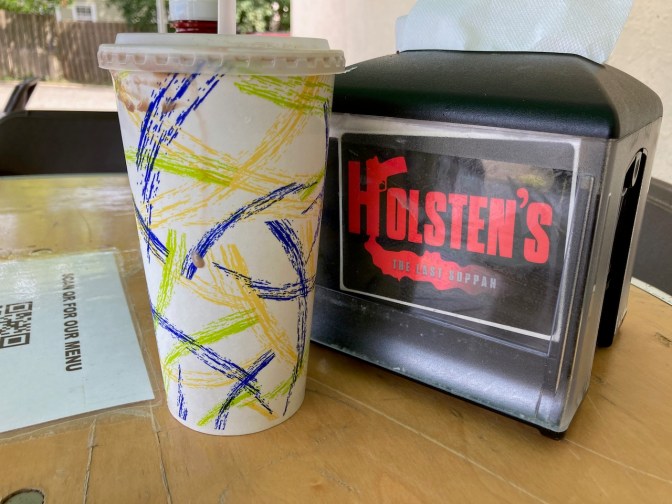 Paper cup beside napkin holder that says HOLSTEN'S THE LAST SUPPAH.