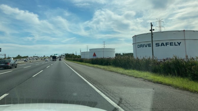 View of New Jersey Turnpike with oil refinery on right, one of which says DRIVE SAFELY.