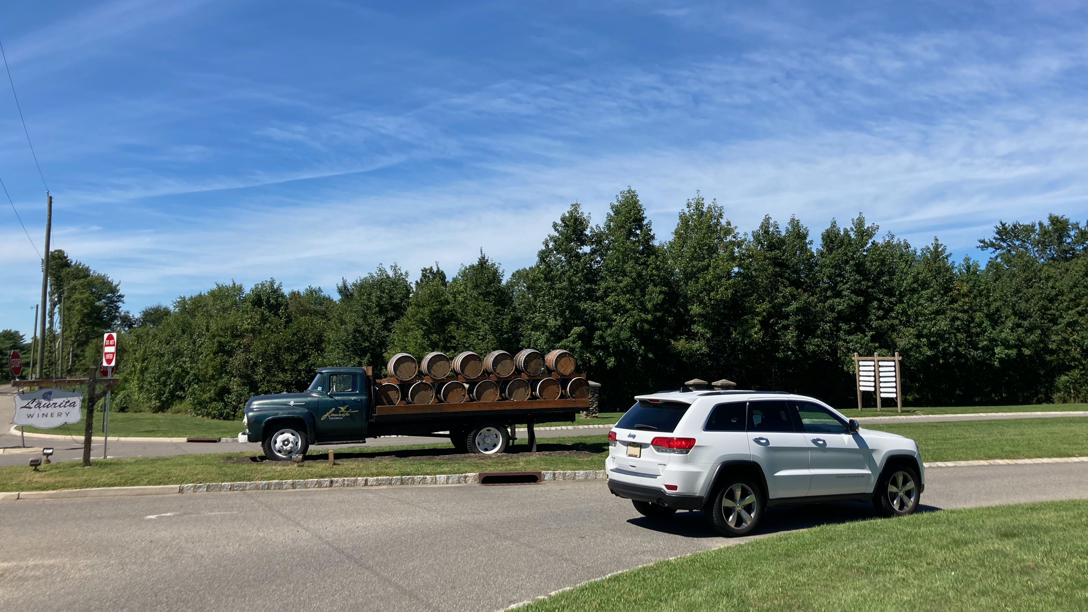 White Jeep Grand Cherokee parked on side of road, with old truck carrying wine barrels in background.
