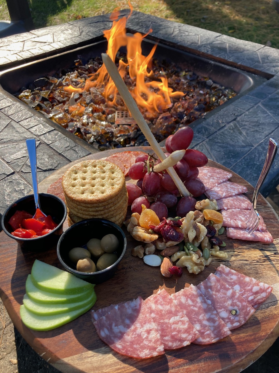 Cheese and fruit spread on wooden platter beside fire pit.