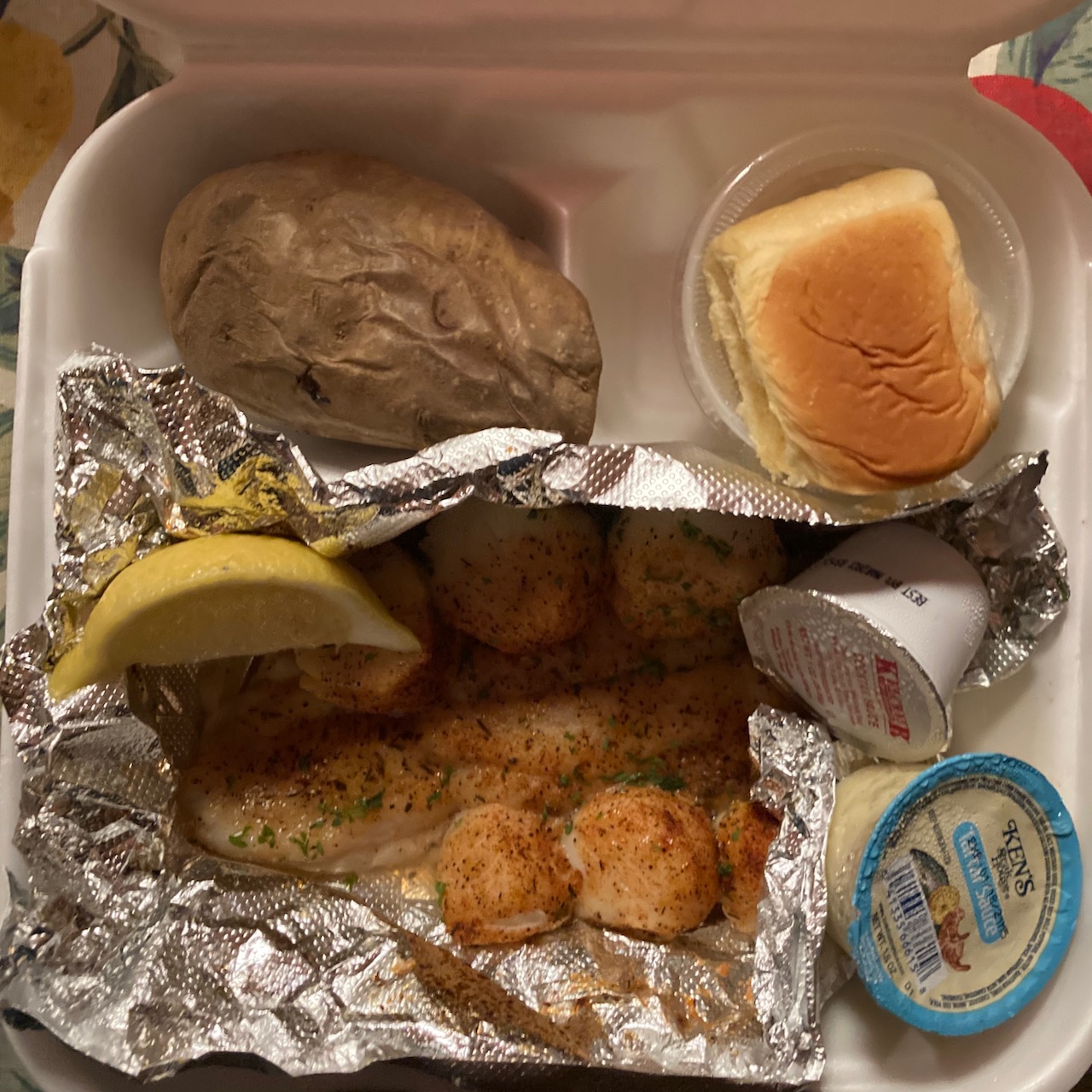 Styrofoam container with roll, baked potato, grouper, scallops, lemon, and sauces.