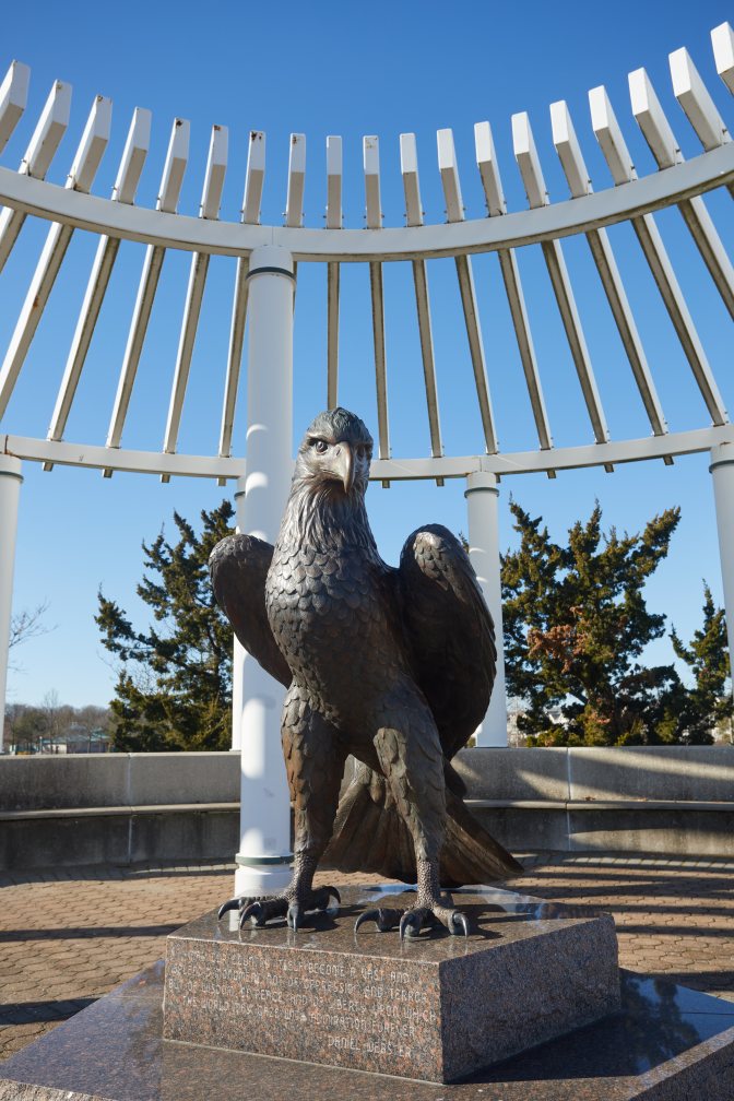 Statue of eagle on pedestal, with white iron structure behind it.