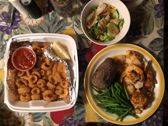 Table with container of calamari, bowl of salad, and a plate with baked potato, green beans, broiled fish, shrimp, and scallops.