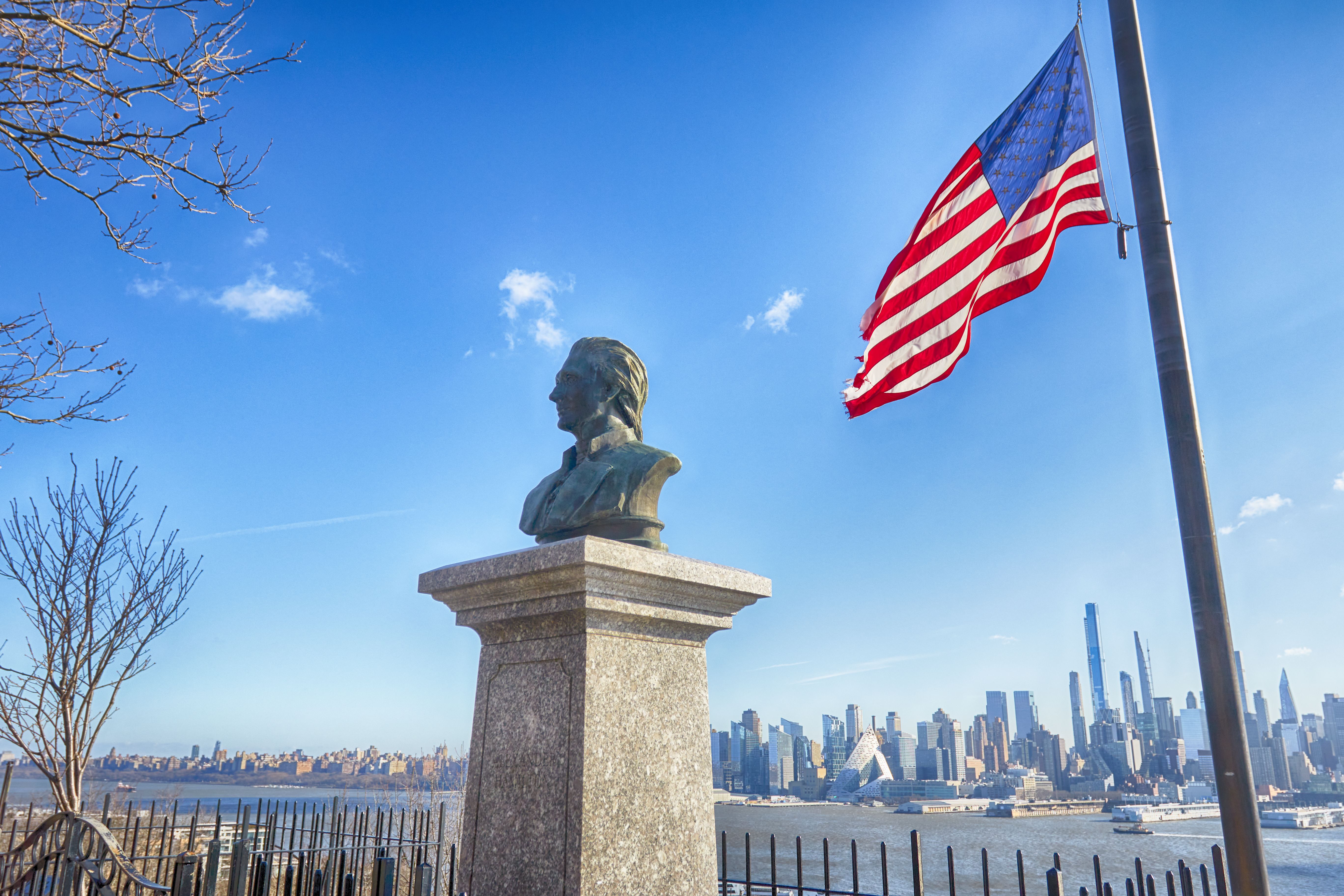 Bust of Alexander Hamilton on plinth, with an American flag at half-staff beside it. Manhattan is visible in the distance.