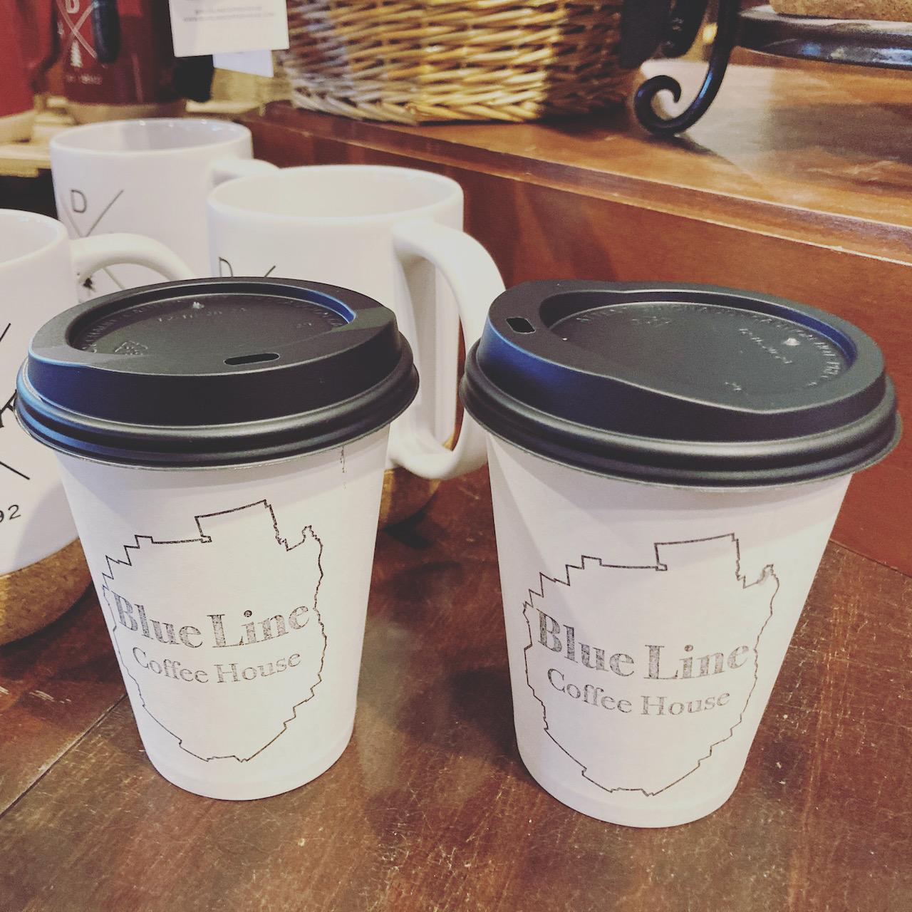 Two paper coffee cups on wooden tables each cup says BLUE LINE COFFEE HOUSE.