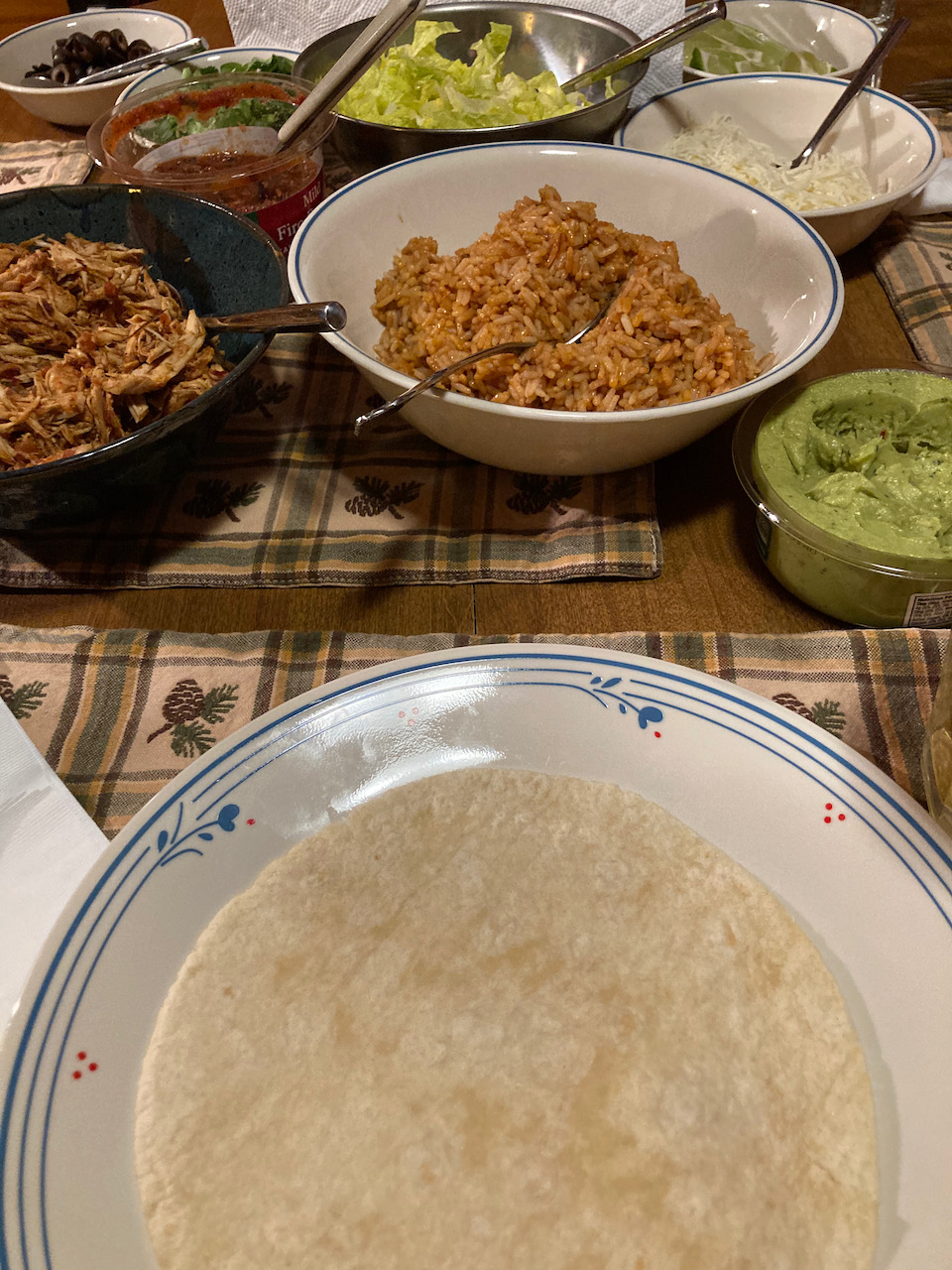 Table with Mexican rice, tortillas, guacamole, shredded chicken, salsa, cheese, lettuce, and black olives.