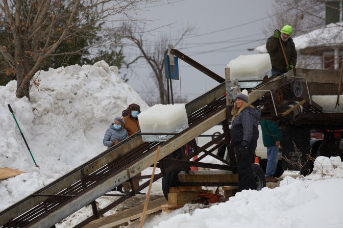 Workers gathered around conveyer belt bringing ice into truck along shore.